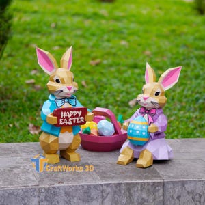 May include: Two papercraft Easter bunnies. One holds a sign saying "Happy Easter", the other holds a decorated egg. A basket of colourful eggs sits between them. The bunnies are tan, with pink ears and bow ties. The bunny on the left wears a blue jacket.