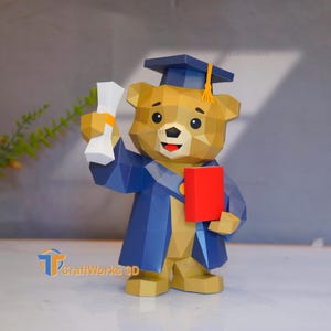 May include: A papercraft graduation bear figurine in shades of tan, blue, and red. The bear wears a graduation cap and gown, holding a diploma and a red book. The CraftWorks 3D logo is visible.