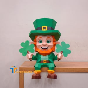 Könnte beinhalten: Eine Papier-Leprechaun-Figur mit grünem Hut, Jacke und Hose, rotem Bart und zwei grünen Kleeblättern. Die Figur sitzt auf einer Holzoberfläche. Das CraftWorks 3D-Logo ist sichtbar.
