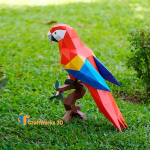 Könnte beinhalten: Ein lebendiger, geometrischer Papagei aus Papier in Rot, Gelb und Blau, der auf einem braunen Ast sitzt. Der Papagei steht vor einem Hintergrund aus grünem Gras und Blättern. Das Logo "CraftWorks 3D" ist sichtbar.