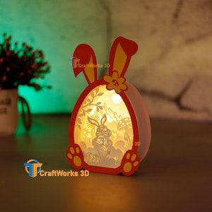 Puede incluir: Una luz nocturna de Pascua de papel en 3D con una cabeza de conejo amarilla y roja y un cuerpo blanco en forma de huevo. El huevo tiene un diseño recortado de un conejo sosteniendo una zanahoria en una escena de bosque. La luz se ilumina desde adentro.