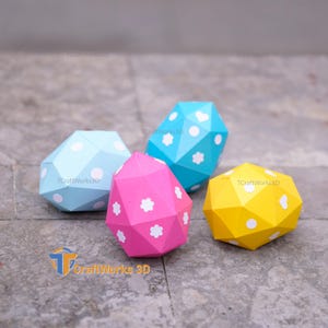 K&ouml;nnte beinhalten: Vier farbenfrohe, geometrische Papierornamente in Blau, Rosa, Gelb und T&uuml;rkis. Jedes hat ein einzigartiges Muster aus wei&szlig;en Punkten und Blumenformen. Die Ornamente werden auf einer grauen Oberfl&auml;che pr&auml;sentiert.