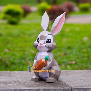 Peut inclure: Figurine géométrique en papier d'un lapin tenant une carotte. Le lapin est principalement gris avec des accents blancs et roses. La carotte est orange avec le dessus vert. La figurine est posée sur une surface en pierre.