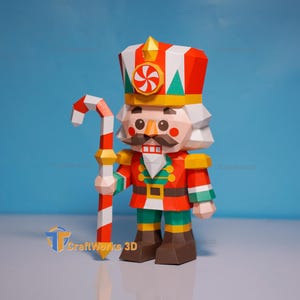 May include: A low-poly papercraft nutcracker figure featuring a red, white, and green uniform with a matching hat adorned with a peppermint candy emblem. The figure holds a red and white striped candy cane staff and has brown boots.