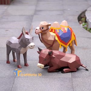 Può includere: Un set di papercraft low-poly composto da un asino grigio, un cammello color sabbia con una sella colorata rossa, blu e gialla, e un toro marrone in posizione di riposo. Il logo CraftWorks 3D è visibile nell'angolo in basso a sinistra.