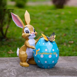 May include: A papercraft Easter bunny figure with a light brown body and pink-lined ears stands next to a large blue egg decorated with white polka dots. A yellow and black butterfly rests on top of the egg. The egg has the text "CraftWorks 3D".