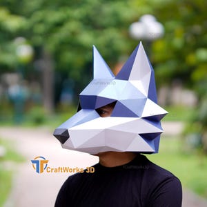 Puede incluir: Una máscara de papel low-poly con forma de cabeza de lobo, que presenta facetas geométricas en tonos azul, blanco y gris oscuro. La máscara tiene orejas puntiagudas, un hocico con la punta de la nariz negra y aberturas para los ojos.