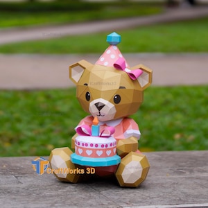 Könnte beinhalten: Papierbastel-Bärenfigur mit Geburtstagskuchen. Der Bär trägt einen rosa Partyhut mit weißen Punkten und eine rosa Schleife. Der Kuchen hat eine blaue Basis, weißes Frosting und rosa Herzdekorationen. Eine brennende Kerze sitzt oben.