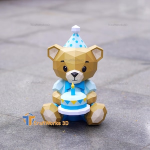 Pode incluir: Um urso de papel castanho com um chapéu de festa azul com bolinhas e uma gravata borboleta azul. O urso está segurando um bolo de aniversário azul e branco com uma vela acesa. O urso é um modelo de papel 3D.