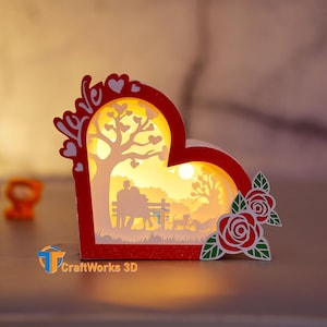 May include: A red heart-shaped wooden box with a 3D scene inside. The scene depicts a couple sitting on a bench under a tree with a full moon in the sky. The box is illuminated from within, creating a warm glow. The words "Love" are carved into the top of the box. The box is decorated with two red roses on the side.