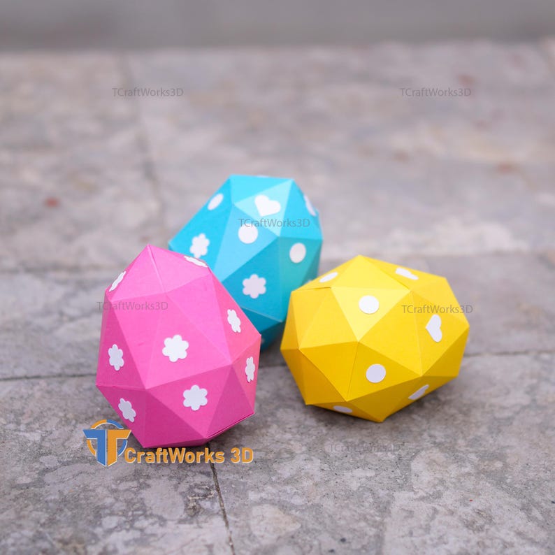 K&ouml;nnte beinhalten: Drei geometrische Papierskulpturen in Rosa, Blau und Gelb. Jede hat ein facettiertes Design und ist mit wei&szlig;en Blumen- oder Punktmustern verziert. Das CraftWorks 3D-Logo ist sichtbar.