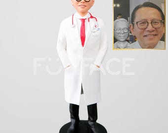 May include: A white doctor figurine with a red tie and stethoscope. The figurine is standing on a black base. A photo of the person the figurine is based on is in the background.