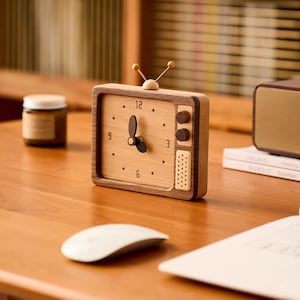 May include: A rectangular wooden analog clock shaped like a vintage television set. The clock face is light wood with dark brown hands and numerals at 3, 6, 9, and 12. It features two small knobs, a speaker grille, and a top antenna detail.