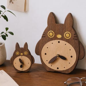 May include: Two wooden wall clocks shaped like a cartoon creature with pointed ears. The clocks feature a dark brown wood exterior, a lighter beige circular clock face, and dark brown hands. The creature design includes yellow eyes and a nose.