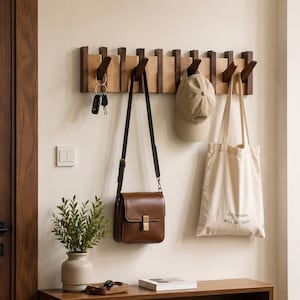 May include: A wall-mounted wooden coat rack featuring a series of vertical slats and angled hooks. The rack is finished in a dark brown wood tone and holds a brown leather shoulder bag, a beige baseball cap, and a cream canvas tote bag.
