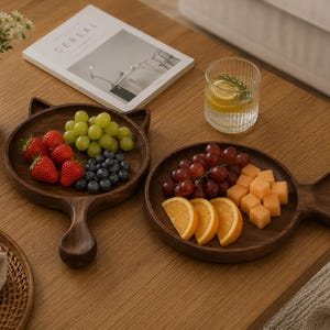 May include: Two dark wood serving boards with handles, one shaped like a cat head and the other like a fish. The cat-shaped board holds strawberries, green grapes, and blueberries. The fish-shaped board holds red grapes, orange slices, and melon cubes.
