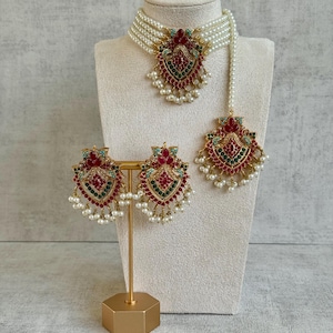 May include: A three-piece jewellery set comprising a choker necklace, earrings, and a headpiece. The set features a gold-toned pendant with red, green, and turquoise accents, and white pearl-like beads. The necklace and headpiece are strung with multiple strands of white beads.