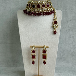 May include: A gold-coloured necklace and earring set. The necklace is a choker with red gemstone accents and dangling beads. The earrings have a matching design with red square and round gemstones. The jewellery is displayed on a white mannequin.