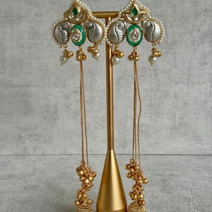 May include: Elaborate gold earrings with detailed craftsmanship. Each earring showcases a silver paisley pattern, green embellishments, and tiny white pearls. Long gold chains with bead clusters and bell-shaped dangles finish the design. The earrings are presented on a gold stand.