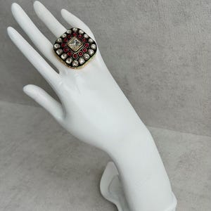 Peut inclure: Une bague carrée avec une pierre centrale transparente, entourée de pierres rouges et blanches, sertie dans un cadre noir et doré. La bague est présentée sur une main blanche sur fond gris.