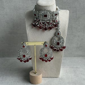 May include: A silver bridal jewellery set comprising a necklace, earrings, and a headpiece. The set features a choker-style necklace with a central square red stone and dangling red beads. The earrings and headpiece echo the necklace design.