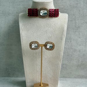 Peut inclure: Un ensemble de bijoux comprenant un collier ras du cou et des boucles d'oreilles assorties. Le collier ras du cou présente une grosse pierre carrée transparente au centre, entourée de petites pierres rouges. Les boucles d'oreilles sont carrées avec une pierre transparente et une bordure dorée.
