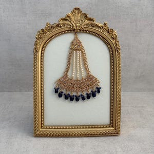 Puede incluir: Un tocado dorado con intrincados detalles de filigrana y perlas, exhibido dentro de un marco dorado decorativo. El tocado presenta un diseño central con cuentas colgantes en forma de lágrima azul marino. El marco tiene un diseño ornamentado de inspiración vintage.