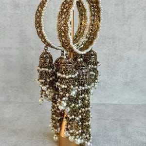 Könnte beinhalten: Goldfarbene Armreifen mit aufwendigen Details und weißen Perlenakzenten. Die Armreifen werden auf einem goldenen Ständer präsentiert, mit baumelnden glockenförmigen Ornamenten, die mit Perlen verziert sind. Der Schmuck hat eine antike Ästhetik.
