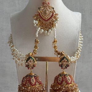 May include: A gold-toned jewelry set featuring a central maang tikka and two matching jhumka earrings connected by strands of white pearls. The pieces are adorned with red, green, and clear crystals, along with small white pearl bead accents.