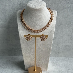 May include: A gold-coloured necklace and matching earrings set displayed on a white bust and a gold stand. The necklace and earrings feature clear, round gemstones set in a delicate design. The jewellery is presented against a neutral grey backdrop.