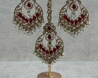 May include: An ornate gold-toned jewelry set featuring a pair of large, teardrop-shaped earrings and a matching headpiece. The earrings and headpiece are adorned with red gemstones, clear crystals, and small white pearls. The set is displayed on a wooden stand.