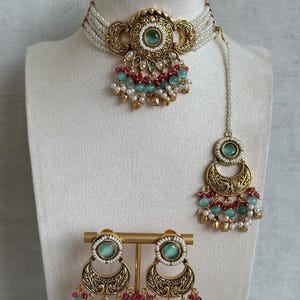 May include: A gold-toned choker necklace featuring multiple strands of white seed beads, a central medallion with a green stone, and dangling red and green beads. Includes a matching maang tikka and a pair of drop earrings with similar motifs.