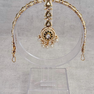 May include: An ornate gold and pearl headpiece with a central pendant and a curved band. The headpiece features white pearls, gold accents, and green and clear gemstone details. The pendant has a cluster of pearls at the bottom.