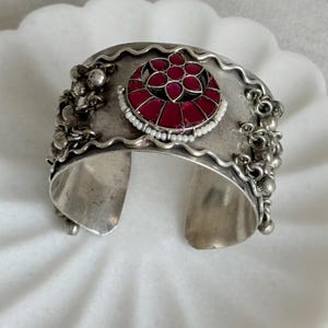 May include: A silver-toned cuff bracelet featuring a central circular medallion with deep red petal-shaped inlays surrounded by a border of small white beads. The edges are wavy, and small silver-toned spherical charms hang from the sides.