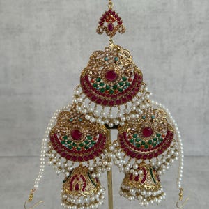 May include: Gold-coloured bridal jewellery set featuring a headpiece and earrings. The set includes ruby-coloured and emerald-green accents, with dangling faux pearls. The earrings have a bell shape. The headpiece has a floral design.