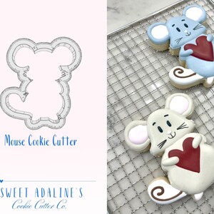 May include: A cookie cutter in the shape of a mouse holding a heart. The cutter is shown next to two decorated cookies, one in light blue and one in beige, both holding hearts.
