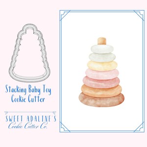 Puede incluir: Un cortador de galletas con forma de juguete apilable para bebés. El cortador está hecho de metal y tiene un acabado liso y brillante. El texto "Stacking Baby Toy Cookie Cutter" está impreso debajo del cortador. El texto "Sweet Adaline's Cookie Cutter Co." está impreso debajo del texto.