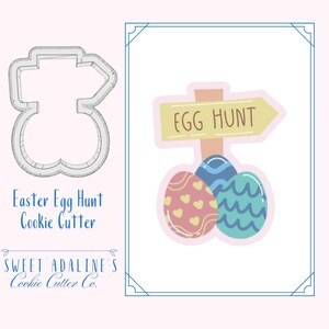 May include: Easter egg hunt cookie cutter with a sign that says "Egg Hunt". The cutter is shaped like three eggs with a sign on top. The sign is yellow with brown text. The eggs are pink, blue, and green with white designs.