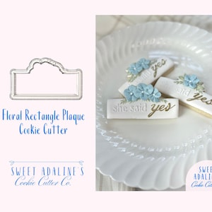 Puede incluir: Un cortador de galletas con forma de placa rectangular floral y galletas decoradas en un plato blanco. Las galletas son rectangulares con las palabras "she said yes" en letras doradas y adornos florales azules. La imagen también incluye el logotipo de Sweet Adaline's Cookie Cutter Co.