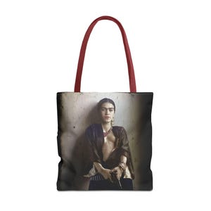 May include: A tote bag with a red handle and a printed image of Frida Kahlo holding a gun. The image is in black and white and has a vintage feel.