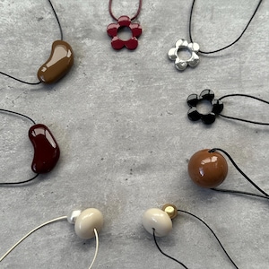 May include: A selection of necklaces featuring diverse pendant designs. These include flower shapes in red, silver, and black, alongside bean and sphere shapes in brown and white. The necklaces are strung on black or white cords.