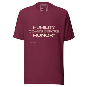 Peut inclure: Un t-shirt bordeaux avec l'inscription "HUMILITY COMES BEFORE HONOR" en beige clair. Le texte "PR 15:33" est en dessous de la citation. Le t-shirt a un col rond et des manches courtes.