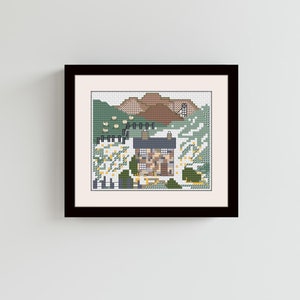 Puede incluir: Una obra de arte de punto de cruz enmarcada que representa una pequeña cabaña ubicada en una ladera verde con colinas marrones en el fondo. La cabaña tiene un techo azul y ventanas blancas. La escena está rodeada de flores blancas.