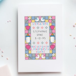 Op de afbeelding: Een ingelijste kruissteek geboorteaankondiging met de naam "Stephanie Jane" en de datum "3-10-95". Het ontwerp bevat babyflessen, harten en het alfabet. Het kleurenschema is pastelroze, blauw, geel en grijs.