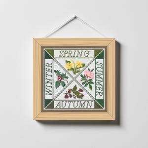 May include: A cross-stitch artwork in a wooden frame, showcasing the four seasons. The design includes floral and botanical motifs, with the words "Spring," "Summer," "Autumn," and "Winter" arranged in a square. The piece is hung by a white string.