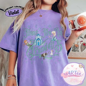 Pode incluir: Camiseta Comfort Colors violeta com a frase "Meet Me In Arendelle" em verde, acompanhada de ilustra&ccedil;&otilde;es de personagens e um castelo. A camiseta tem um corte folgado e gola redonda.