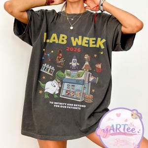 Op de afbeelding: Donkergrijs T-shirt met de tekst "LAB WEEK 2026" in geel. Het shirt heeft een kleurrijke afbeelding van laboratoriumapparatuur, een dinosaurus in een labjas en de zin "TO INFINITY AND BEYOND FOR OUR PATIENTS".