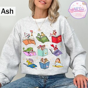 Puede incluir: Sudadera gris ceniza con ilustraciones coloridas de personajes animados leyendo libros. El dise&ntilde;o incluye personajes como un d&aacute;lmata, ranas, un drag&oacute;n y otros, cada uno con un libro abierto. La sudadera tiene cuello redondo y mangas largas.