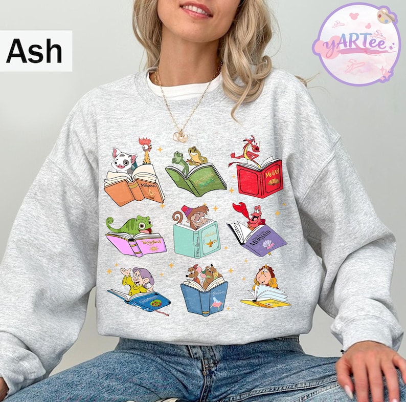 Puede incluir: Sudadera gris ceniza con ilustraciones coloridas de personajes animados leyendo libros. El dise&ntilde;o incluye personajes como un d&aacute;lmata, ranas, un drag&oacute;n y otros, cada uno con un libro abierto. La sudadera tiene cuello redondo y mangas largas.