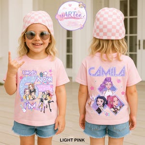 May include: A light pink t-shirt with the text "I WAS 5 NOW I'M 6" and a graphic of cartoon characters. The back of the shirt has the name "CAMILA" and character graphics. The child is wearing a pink and white checkered beanie and blue jean shorts.